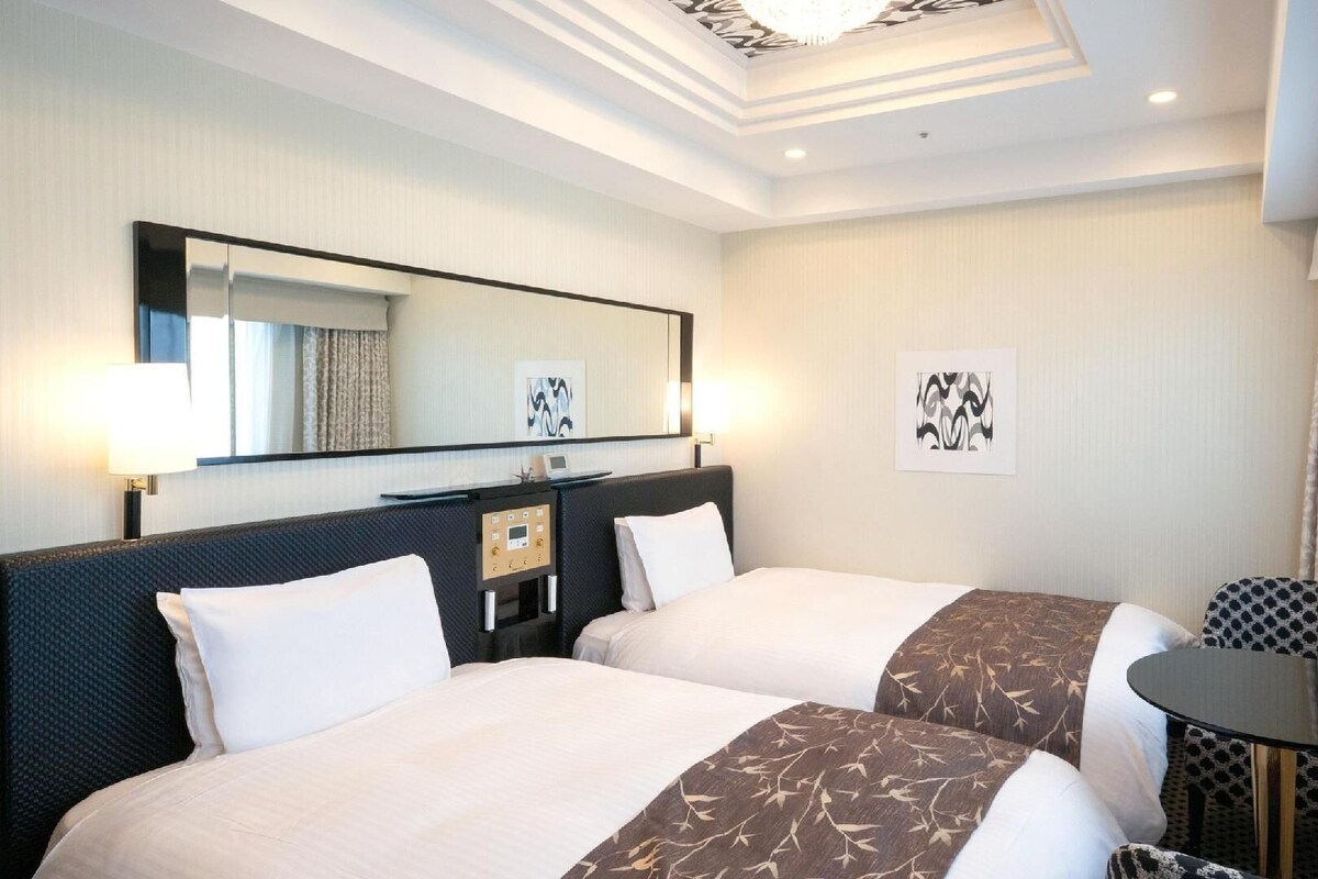 A hotel room with two beds, a desk, a chair, a small table, a mirror, and artwork on the walls.
