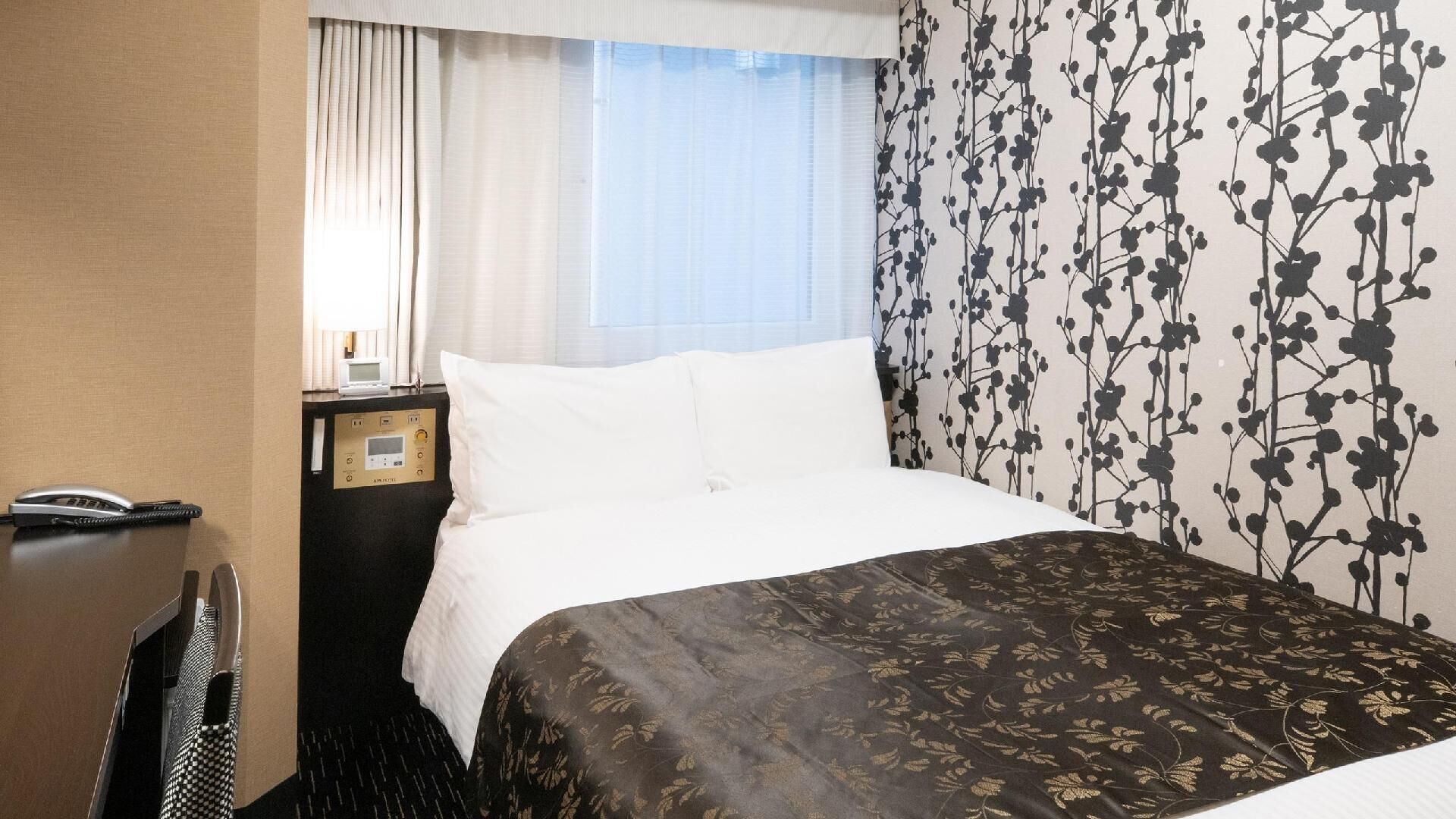 Ett hotellrum med en säng, ett nattduksbord med en telefon och en kontrollpanel, ett fönster med gardiner och en vägg med ett blommönster.