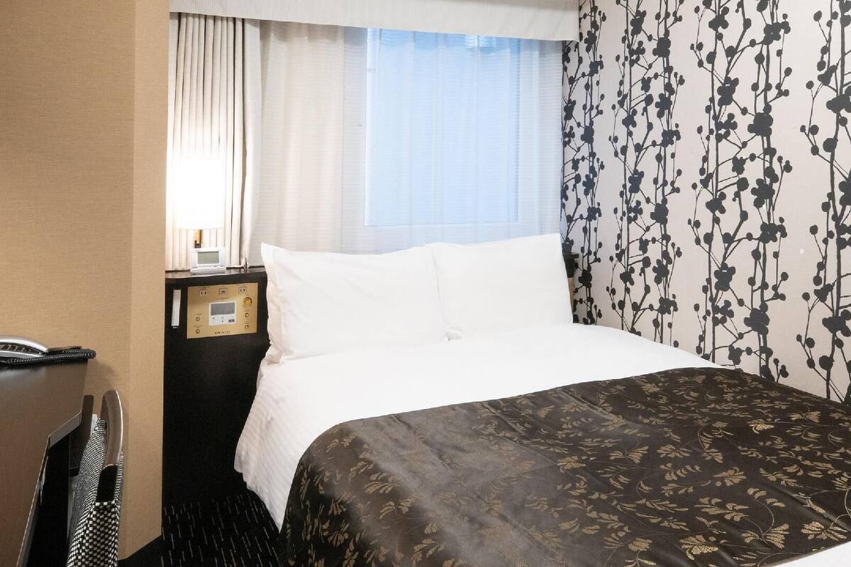 A hotel room with a bed, a bedside table with a phone and a control panel, a window with curtains, and a wall with a floral pattern.