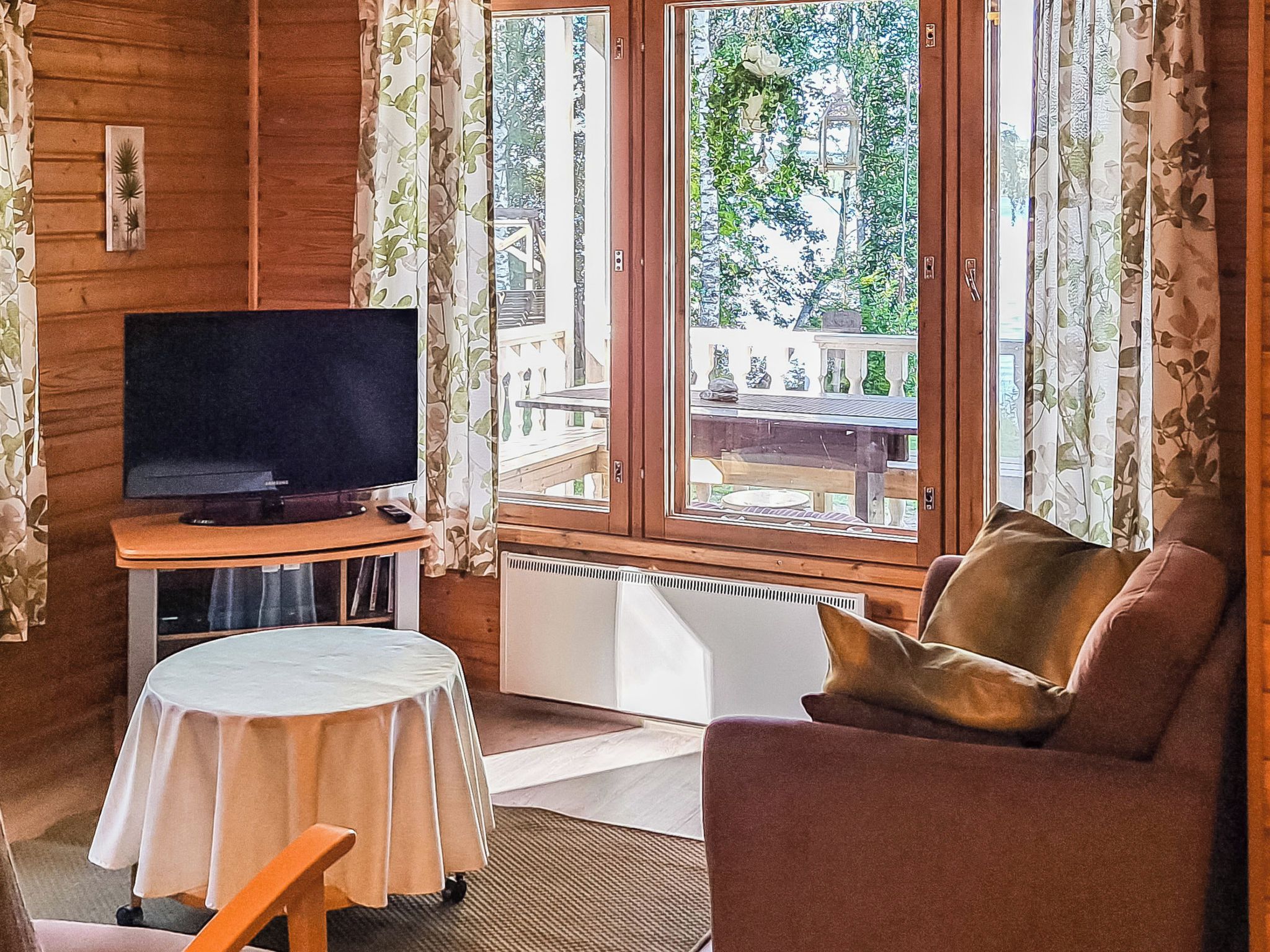 Ein Zimmer mit Fernseher auf einem Schrank, einer Couch, einem kleinen Tisch mit Tischdecke und einem Fenster mit Vorhängen.