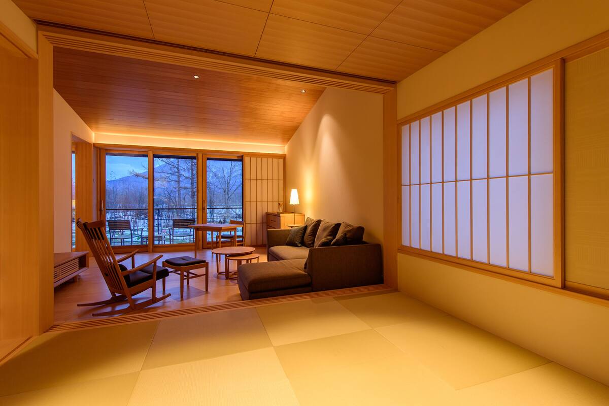 Perinteinen japanilaistyylinen huone, jossa on tatamimatto, matala puukatto, liukuvat shoji-seinäpaneelit, sohva, keinutuoli ja pieni pöytä.
