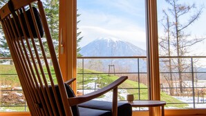 Una silla de madera con cojín y una mesita pequeña, ubicada frente a una ventana que ofrece vistas de un paisaje nevado y una montaña.