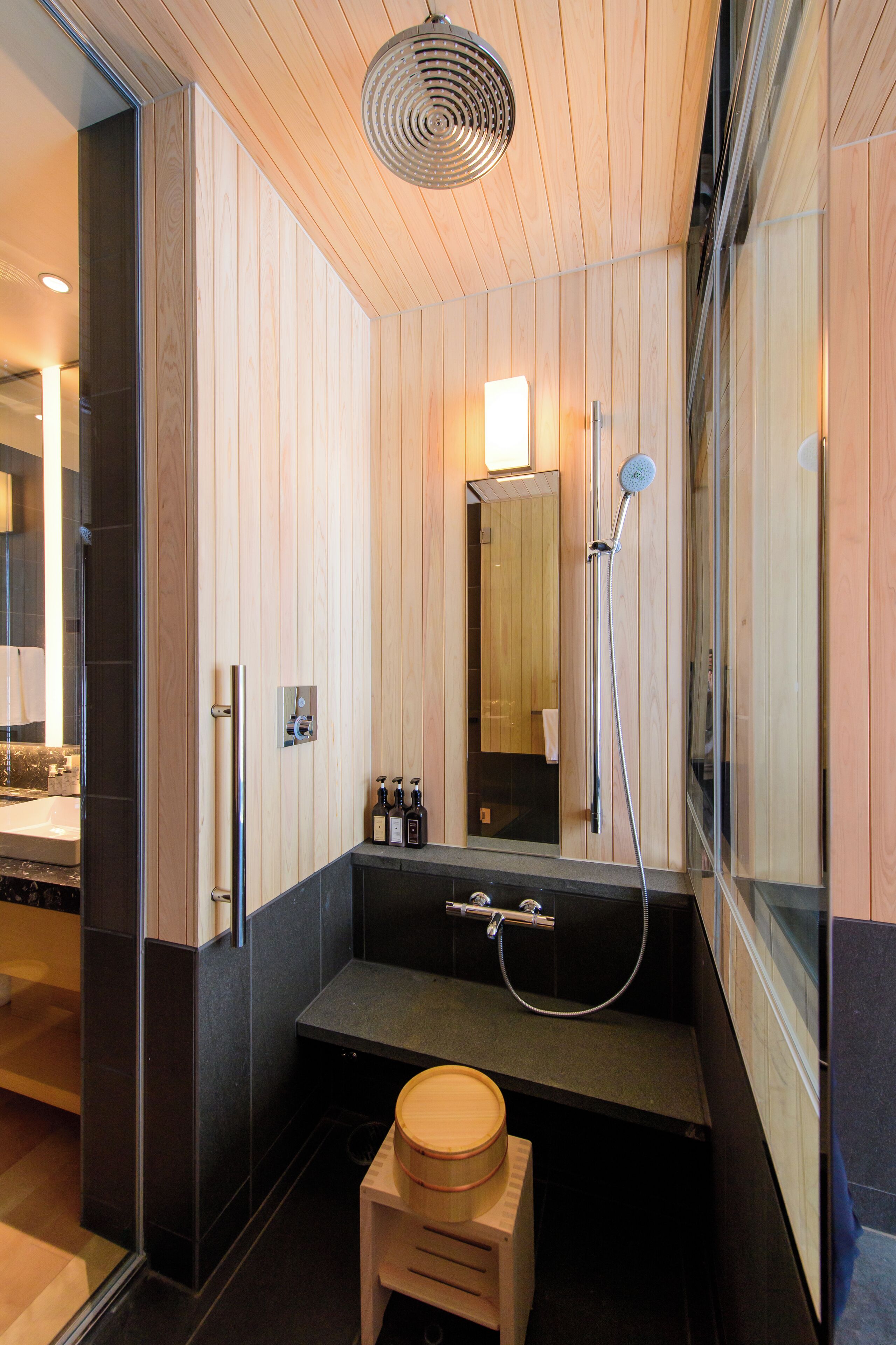 A modern bathroom with wooden panel walls, a black sink, a wooden stool, a handheld showerhead, and a mirror.