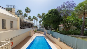 En swimmingpool med blå bund, omgivet af et hvidt hegn, palmer og en bygning med beige facade.
