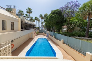A swimming pool with a blue interior, surrounded by a white fence, palm trees, and a building with a beige facade.
