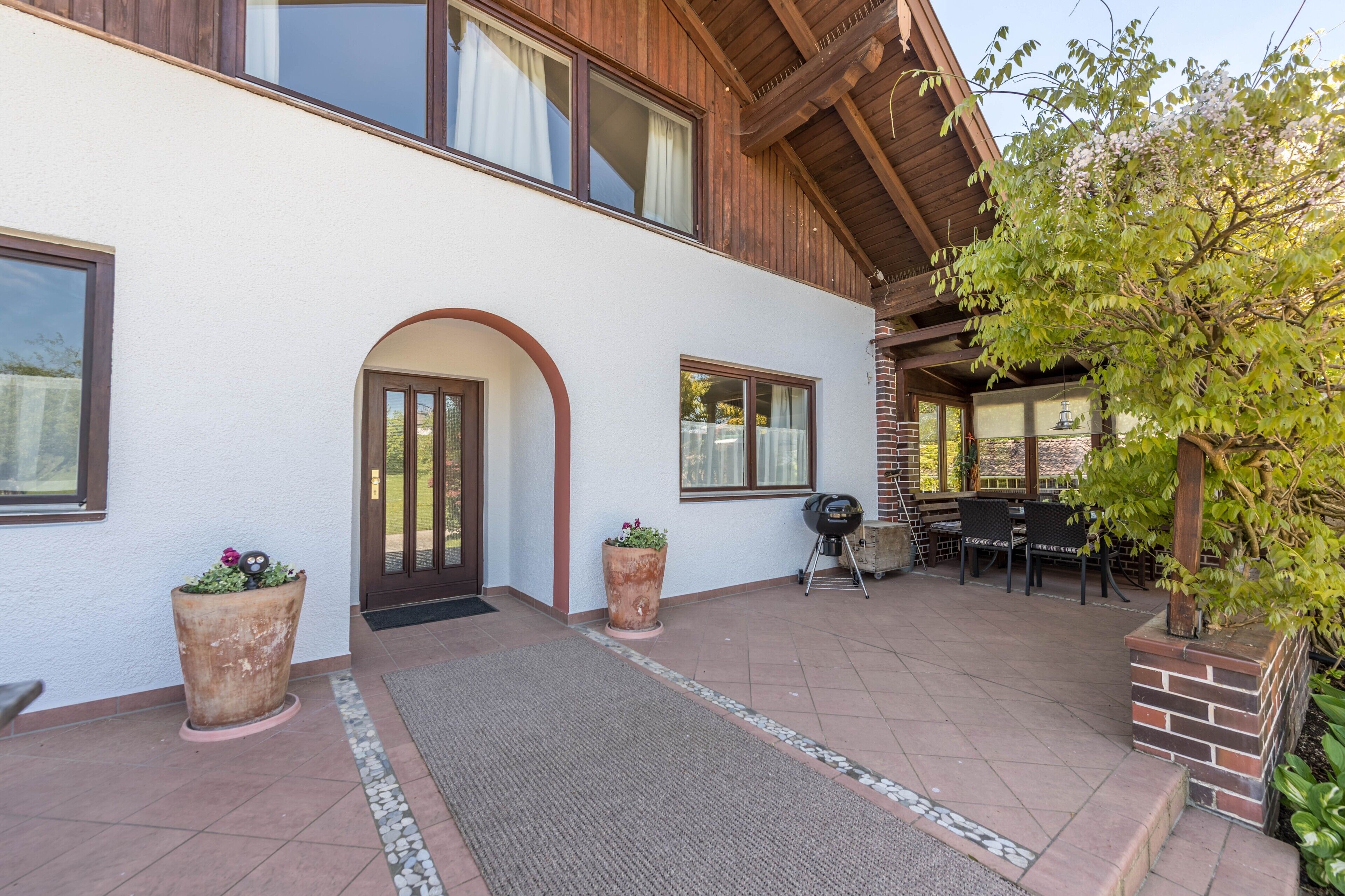 Ein Gebäude mit weißer Fassade, Holzbalken, einer überdachten Terrasse mit Sitzgelegenheiten und Topfpflanzen.