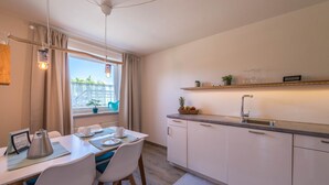A modern kitchen with a dining area, a window with curtains, and a shelf with kitchenware. - Apartment "Julia" with Lake View, Mountain View, Wi-Fi, Garden & Terrace (Taching am See)