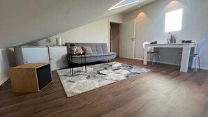 Ett modernt sovrum i loftstil med ett sluttande tak, en grå soffa, ett litet bord, ett skrivbord och ett träskåp.