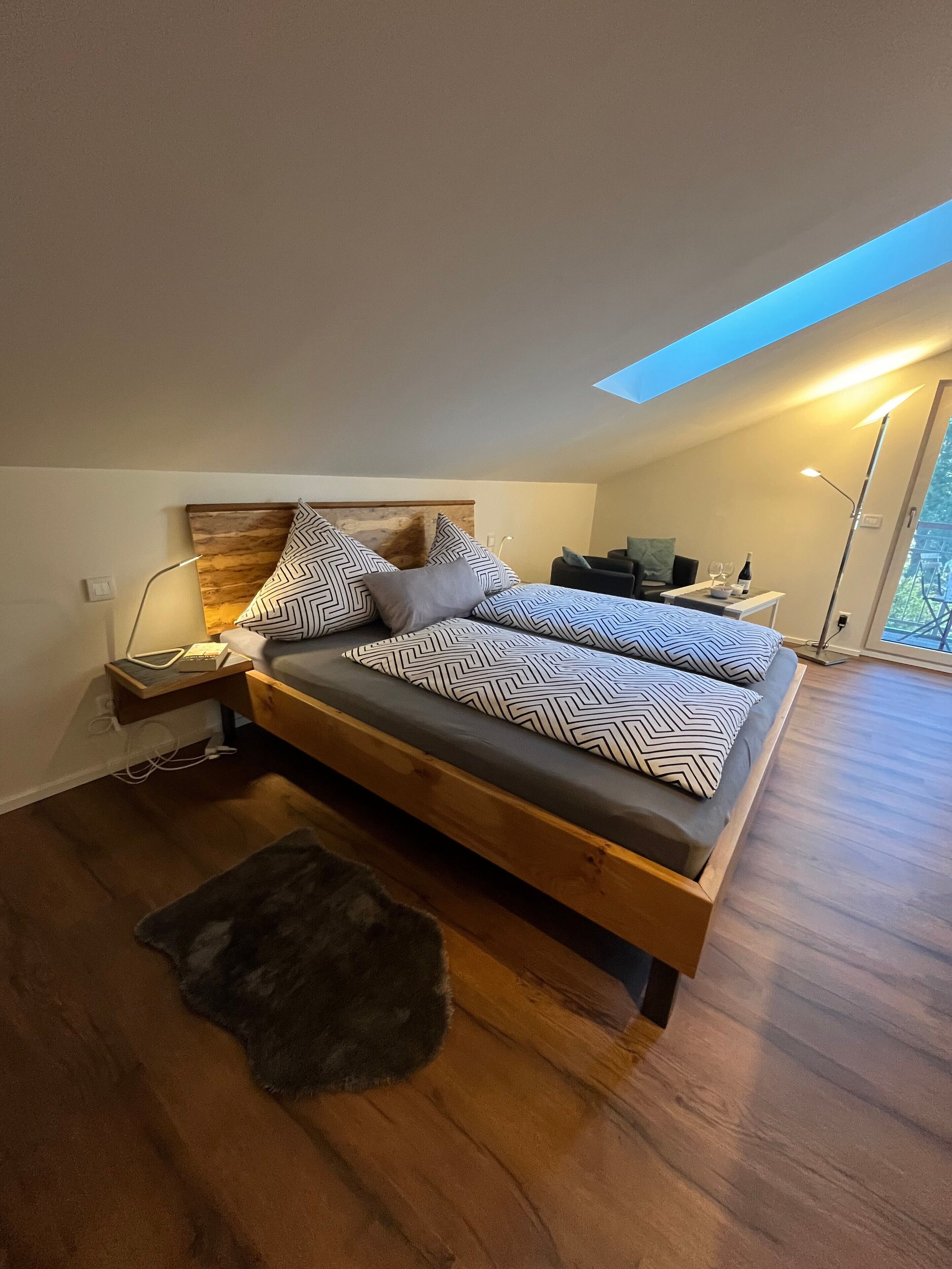 A modern bedroom with a wooden bed, patterned pillows, a nightstand, a desk with a bottle and glasses, and a skylight.