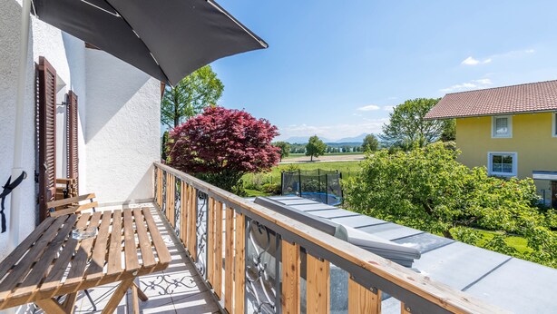Un balcon doté d’une table en bois, d’une rambarde en métal et offrant une vue sur un paysage verdoyant, avec des arbres et un bâtiment.