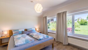 Een slaapkamer met een groot bed, bedlampen, een raam met gordijnen en uitzicht op groen.