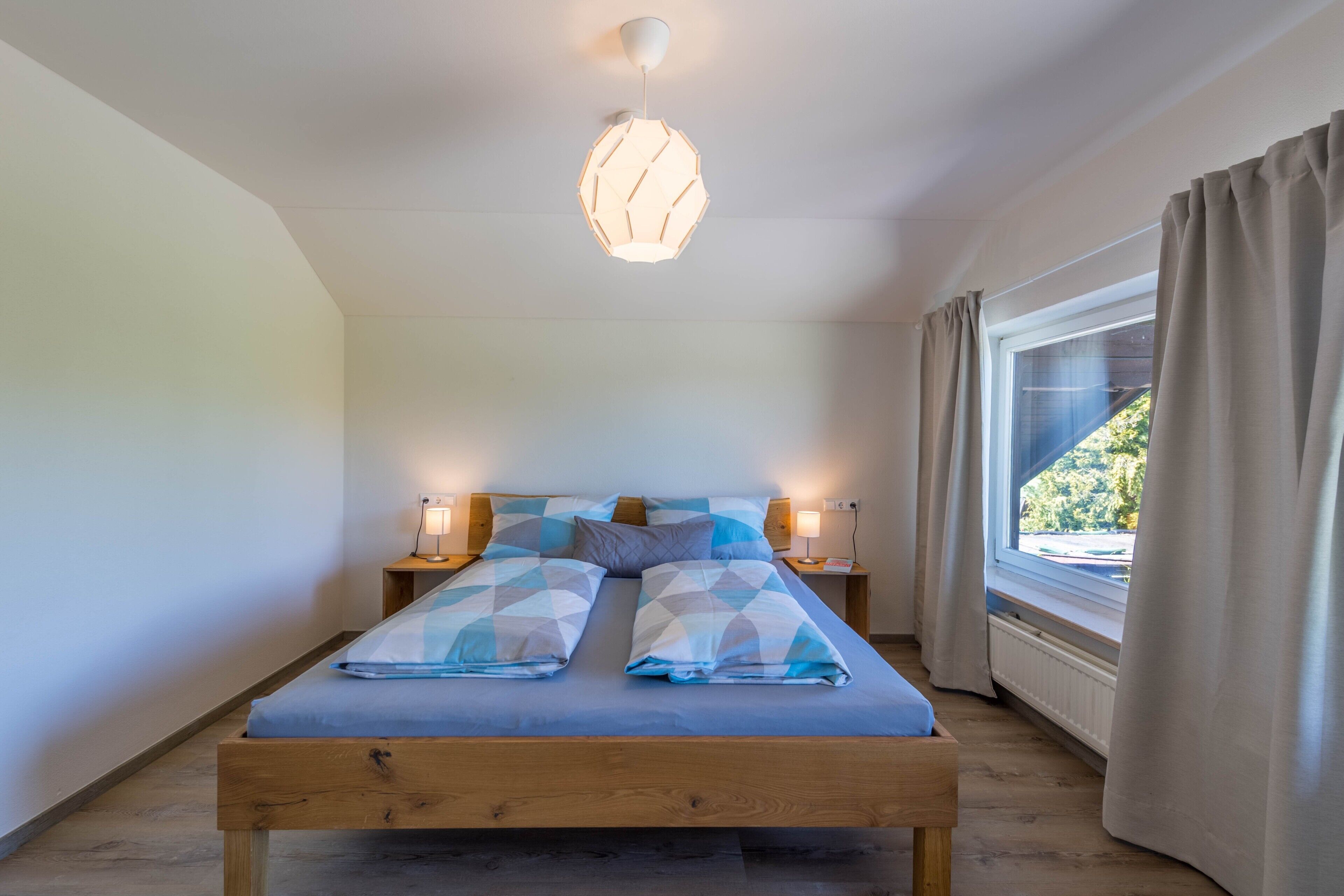 Ein Schlafzimmer mit einem hölzernen Bett, Nachttischlampen, einem Fenster mit Vorhängen und einer Deckenleuchte.