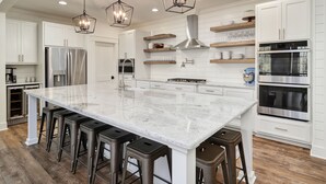 A modern kitchen with a large island, bar stools, stainless steel appliances, and wooden shelves.
