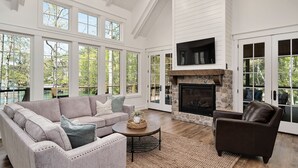 A spacious living room with a stone fireplace, large windows, and a high ceiling.