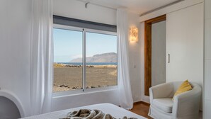 A bedroom with a large window, a bed, a chair, and a view of a beach and mountains. - Modern House "El Loft" with Sea and Mountain View, Wi-Fi & Terrace (Famara)