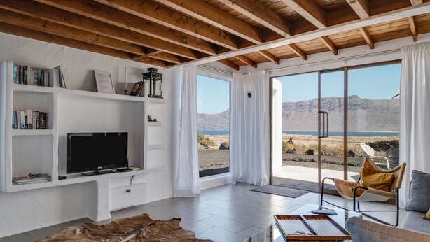 A modern living room with a television, bookshelves, a glass coffee table, and a view of the desert landscape. - Modern House "El Loft" with Sea and Mountain View, Wi-Fi & Terrace (Famara)