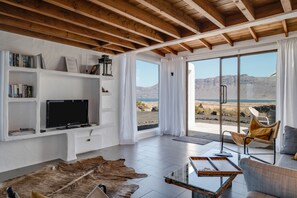 Un salon moderne avec une télévision, des étagères à livres, une table basse en verre et une vue sur le paysage désertique.