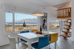 Zona de menjador amb taula, cadires, ampolla i plat de fruita. Una finestra gran ofereix vistes a un paisatge desèrtic.