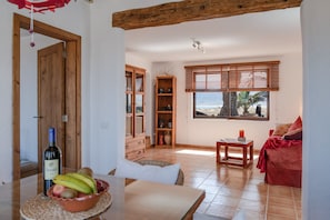 Ein Zimmer mit einer Holztür, einem Tisch mit Flasche und Obst, einem Korbstuhl, einem roten Sofa und Meerblick durch ein Fenster.