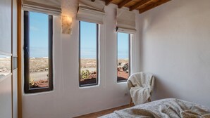 Un dormitorio con vistas a un paisaje desértico a través de amplios ventanales, techo de madera y cama con manta estampada.