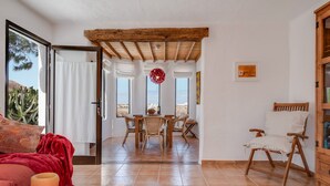 Una sala de estar con techo de madera, un comedor con mesa y sillas, una lámpara colgante roja y vistas a través de un puerta abierta.