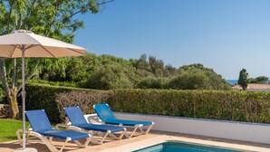 Zona junto a la piscina con sillas de descanso y una sombrilla, rodeada de vegetación y una vista despejada al mar.