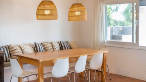 Un comedor con una mesa de madera, sillas blancas, un sofá con cojines estampados y dos lámparas colgantes de mimbre.