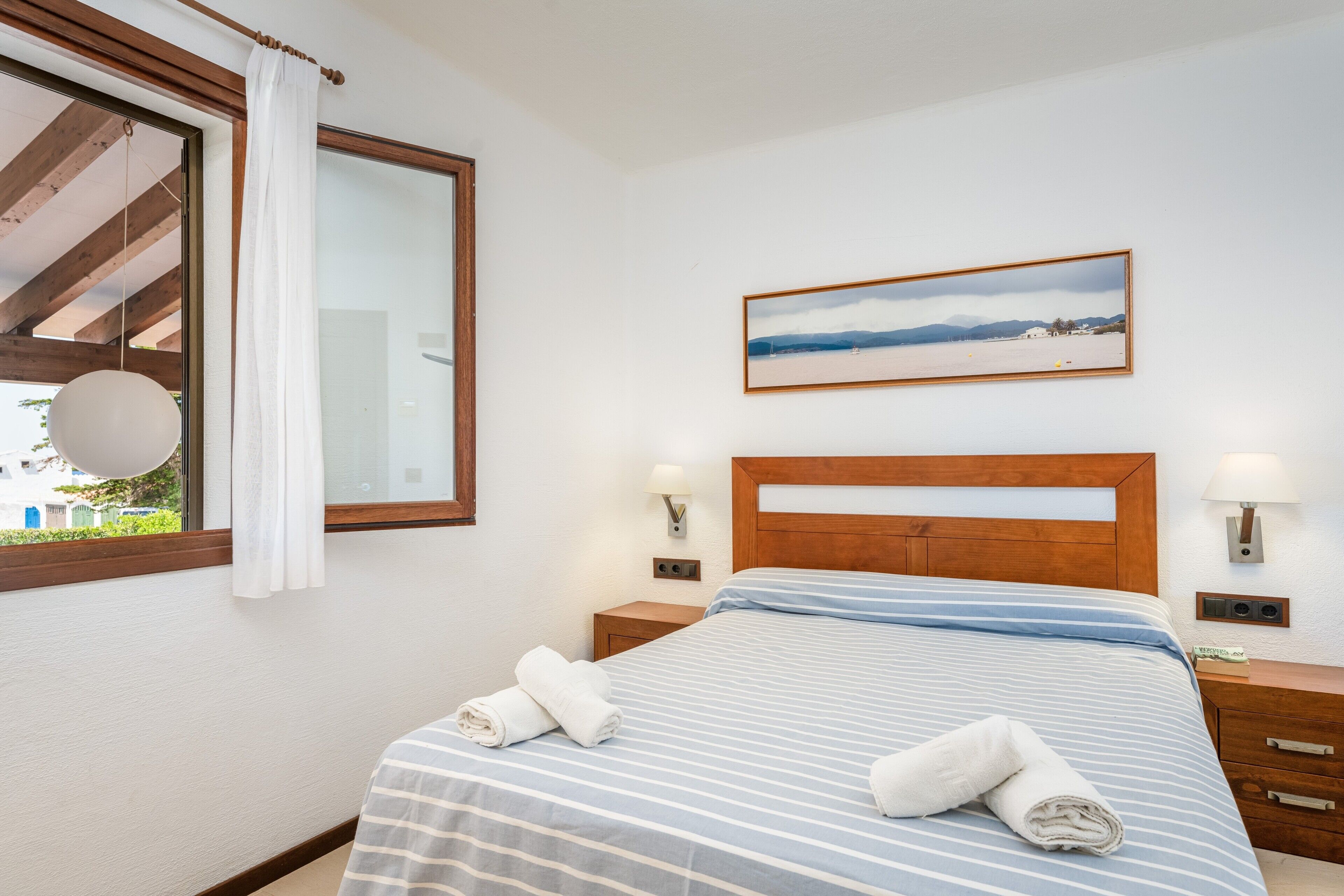 Una camera d'albergo con un letto, una finestra vista, un quadro appeso e un comodino con una lampada.