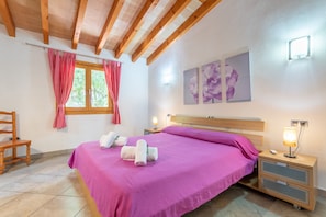 A bedroom with a bed, wooden headboard, pink curtains, a window, a chair, a bedside table with a lamp, and artwork on the wall. - Mediterranean Finca "Es Puch" with Mountain view, Balcony, Garden, Terrace & Pool (BUGER)