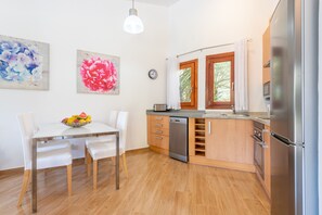 A modern kitchen with a dishwasher, oven, and refrigerator. There is a dining table with chairs and a fruit bowl.