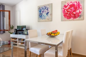 A dining area with a glass table, white chairs, a fruit bowl, and two abstract paintings on the wall.