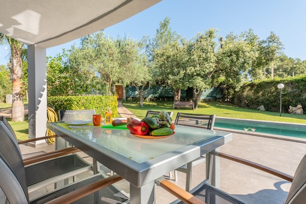 A glass-top table with a bowl of vegetables, outdoor seating, and a view of a swimming pool.
