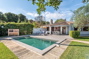A swimming pool with a patio area, a barbecue grill, and a house with a tiled roof.