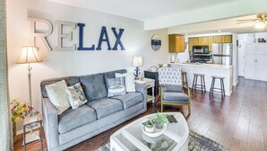 A living room with a grey sofa, a coffee table with plants, a kitchen area with wooden cabinets, and a large 'RELAX' sign on the wall. - 2 Mi to Silver Dollar City: Reed Springs Condo (Reeds Spring)
