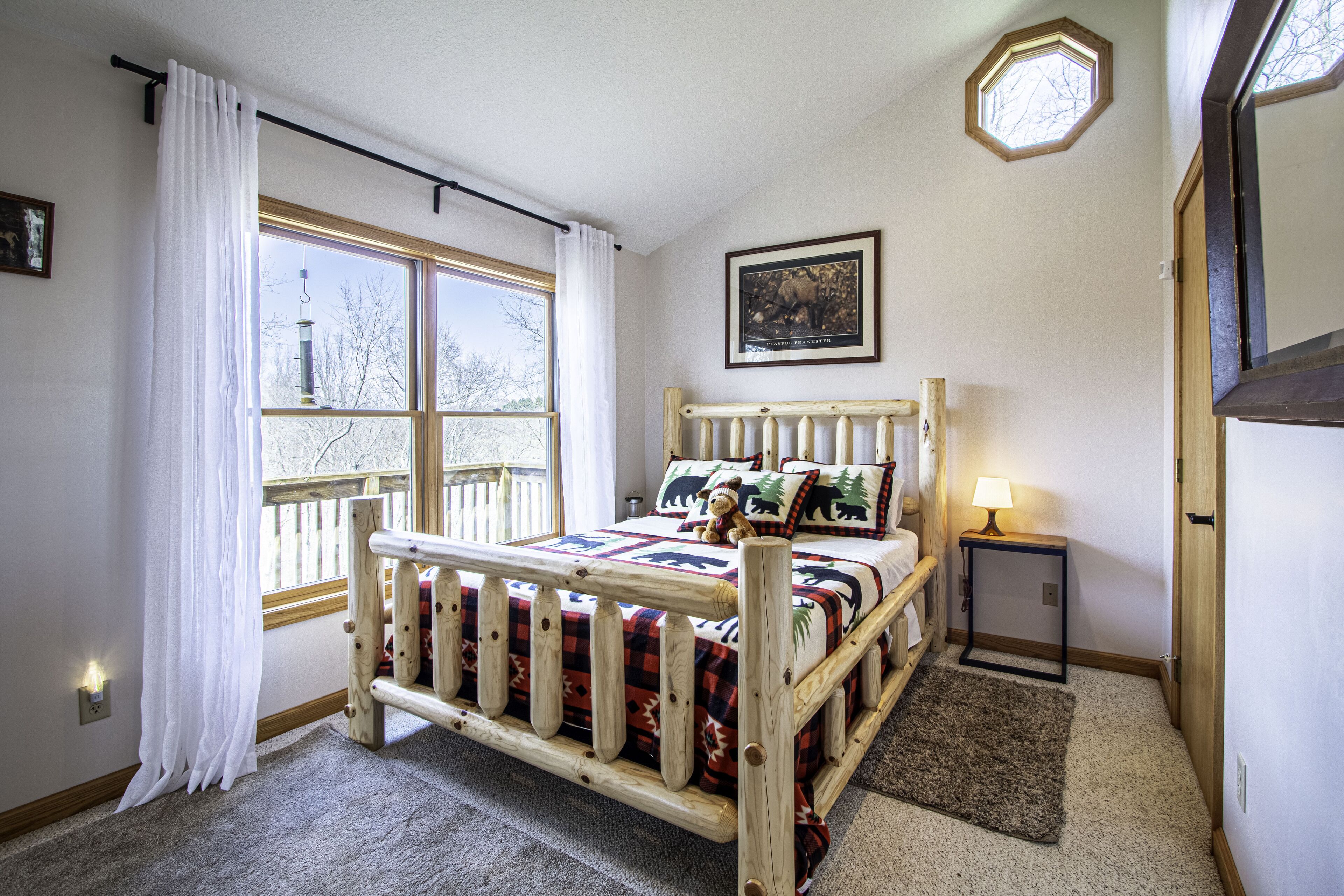 A bedroom with a wooden bed, a window with white curtains, a bedside lamp, and a framed picture on the wall.