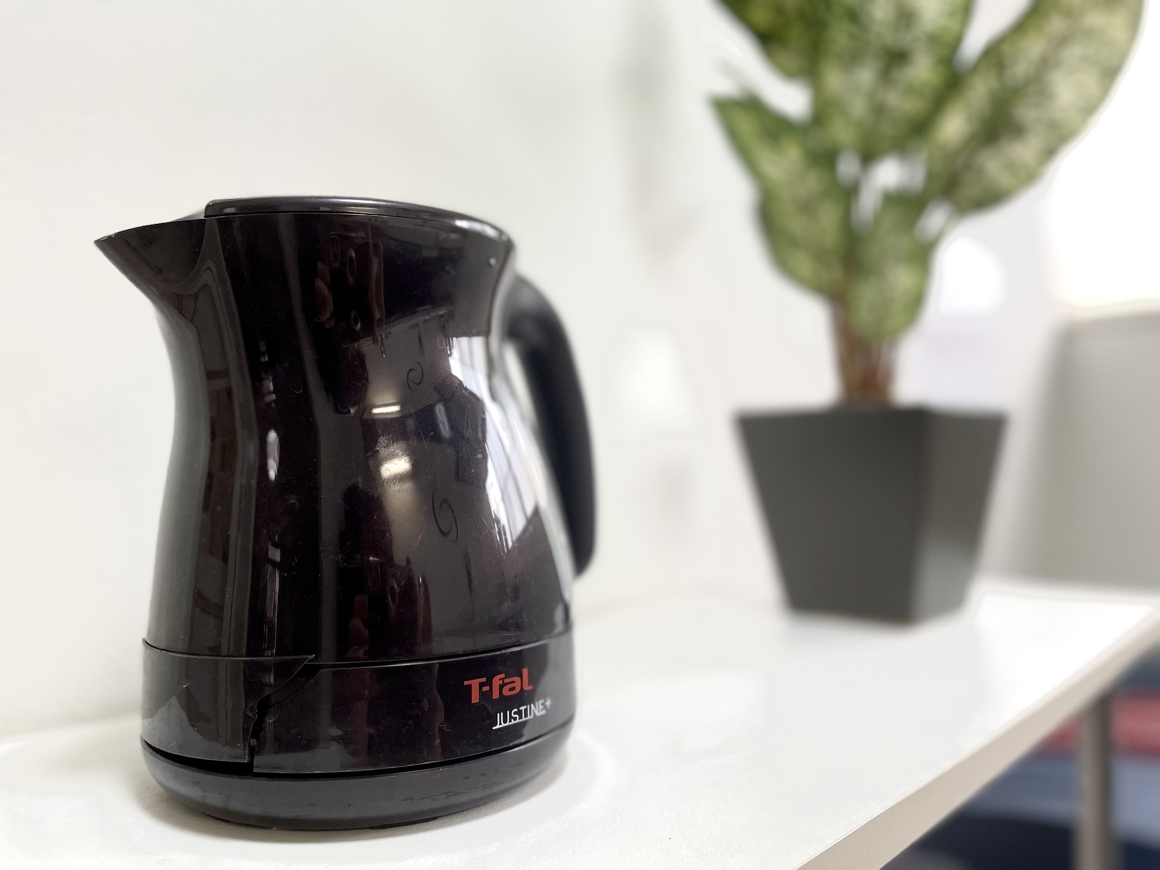 Uma cafeteira Tefal Justine preta sobre uma superfície branca, com uma planta em vaso ao fundo.