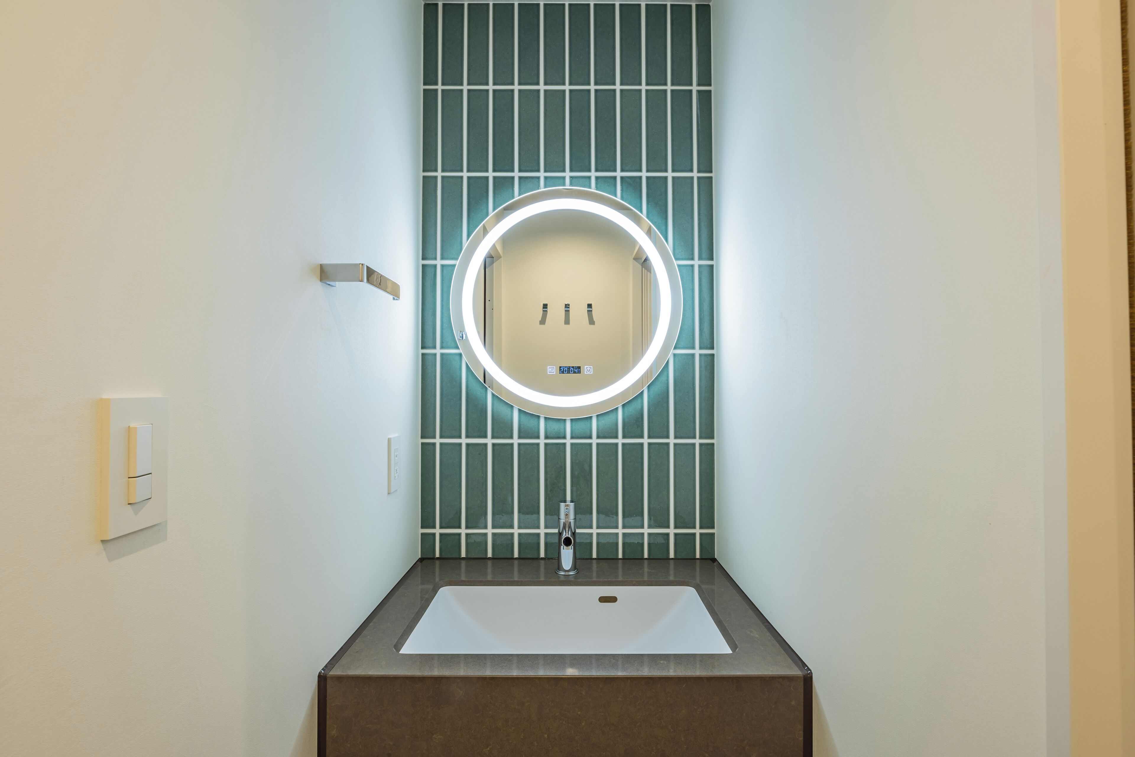 Ein Badezimmer mit einem rechteckigen Waschbecken, einem runden Spiegel mit Beleuchtung und einer gefliesten Wand.