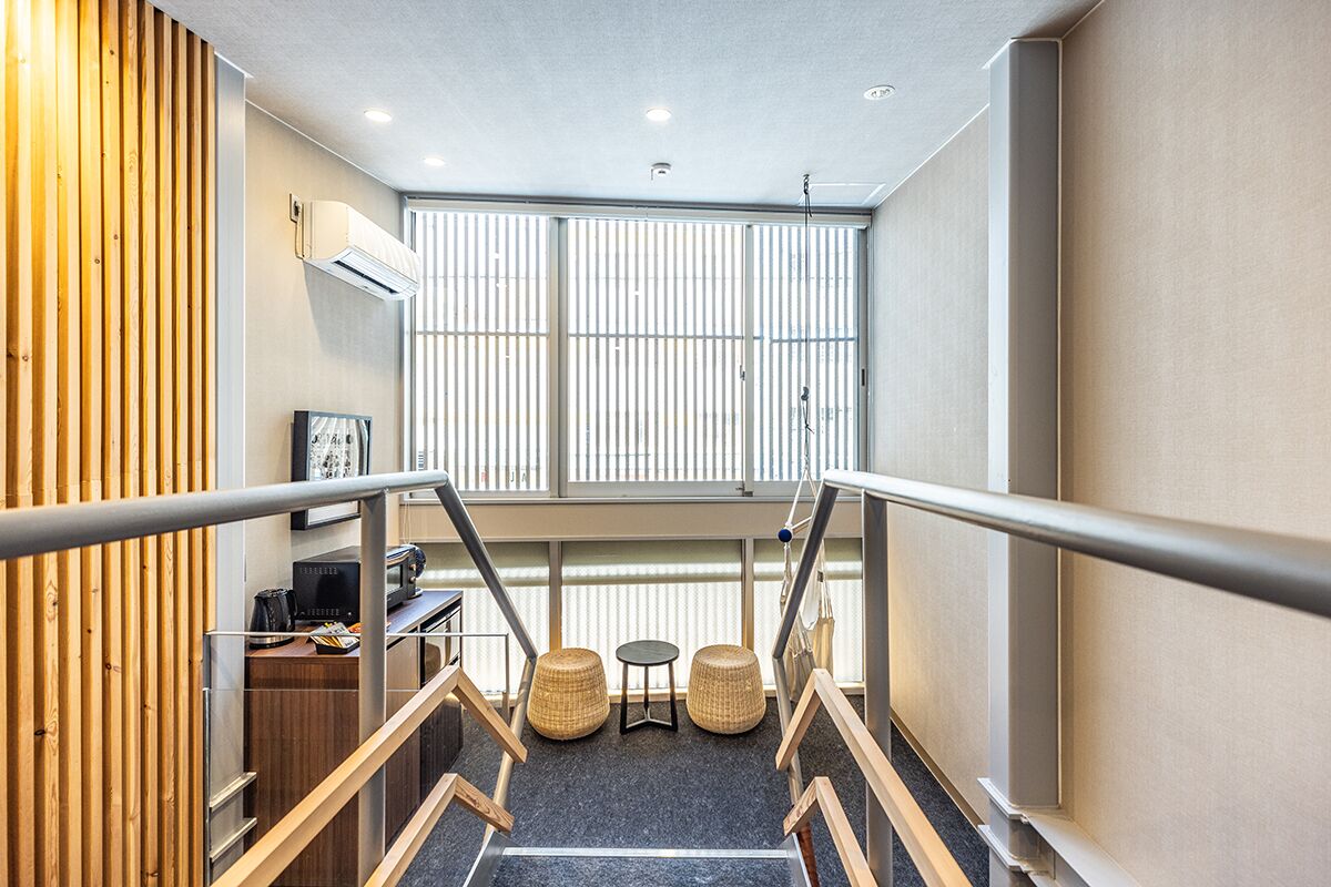 Een modern hotelinterieur met een trap, houten lambrisering, een bureau met een koffieapparaat, twee rieten krukken en een groot raam met jaloezieën.