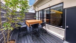 Una terraza de madera con una mesa, sillas, una planta en maceta y una ventana con persianas.