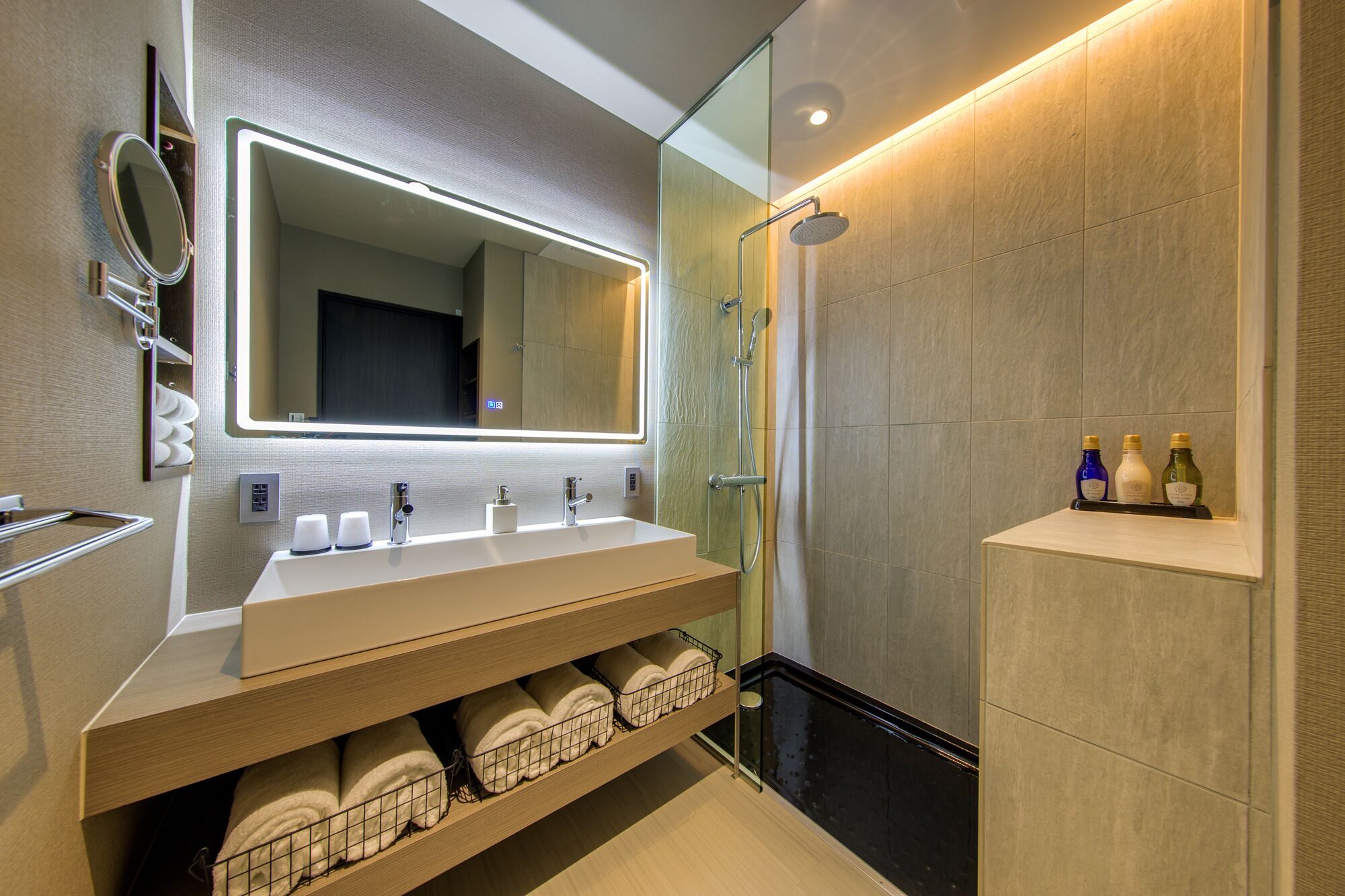 A modern bathroom with a large mirror, two sinks, a shower, and a shelf with toiletries.