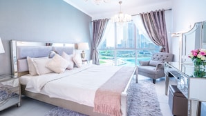 A modern bedroom with a large bed, a sitting area with a chair, a vanity with a mirror, and a view of a cityscape through large windows. - Elite Royal Apartment - Burj Khalifa & Fountain view - Opal (Dubai)
