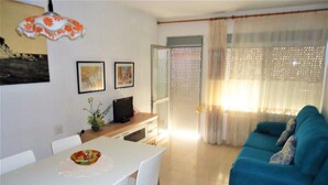 A living room with a blue sofa, a dining table with chairs, a TV on a wooden cabinet, and a window with sheer curtains. - Les Doyes Bl 3 1-36  LEK (Peñiscola)