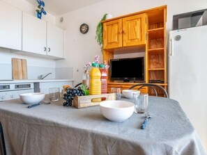 A kitchen with white cabinets, a wooden cabinet with a TV, and a dining table set with bowls, glasses, and a fruit basket. - Apartment 3 Mats by Interhome (Canet-Plage)