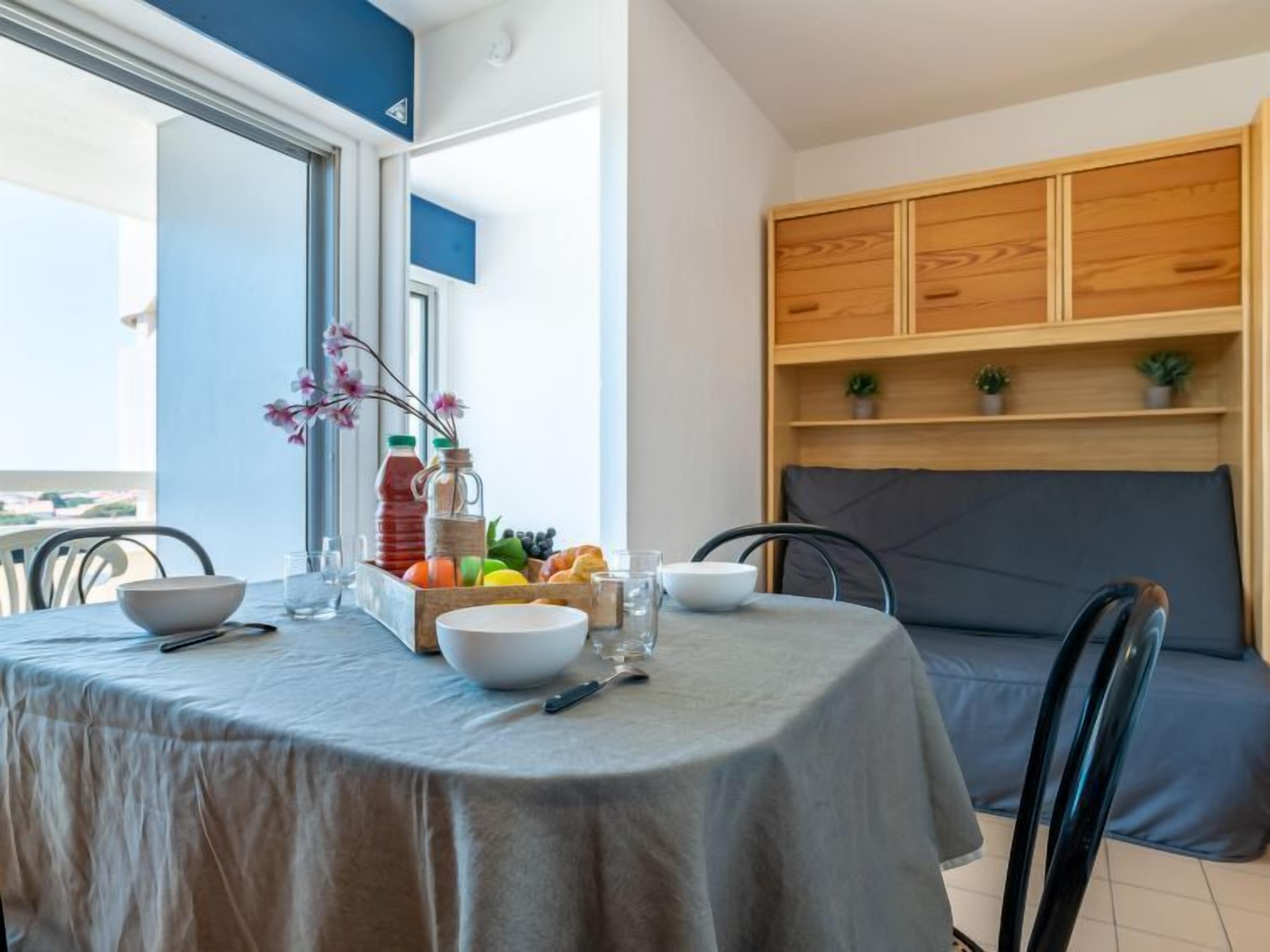 Un comedor con una mesa preparada para dos personas, una cesta de frutas y una botella de agua.