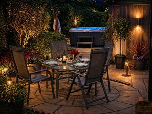 Un patio al aire libre con una mesa lista para comer, rodeado de sillas, plantas en macetas y una bañera de hidromasaje al fondo.