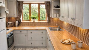 A kitchen with a large window, a sink, a stove, and a countertop with two mugs and a plate of toast. - Measure Cottage - Sleeps up to 4 - Henley in Arden - HOT TUB (Henley-in-Arden)