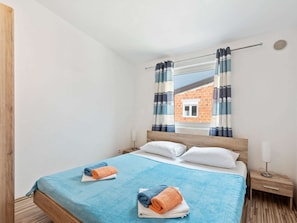 Ein Schlafzimmer mit einem Bett, zwei Nachttischen, einem Fenster mit Vorhängen und Blick auf ein Backsteingebäude.