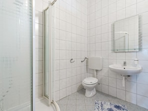 Ein Badezimmer mit weißen Fliesen, einer Duschkabine aus Glas, einer Toilette und einem Waschbecken mit Spiegel darüber.