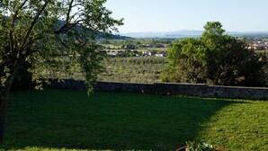 Eine begrünte Außenanlage mit einer Steinmauer, einem Blumentopf und Blick auf ein Dorf und Berge in der Ferne.