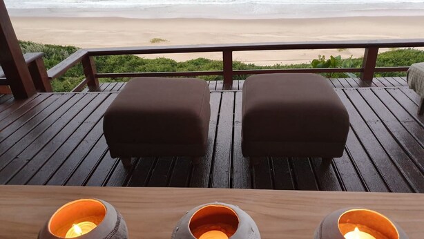 Una terraza de madera con dos sillones acolchados y tres velas encendidas sobre una mesa, con vista a la playa.